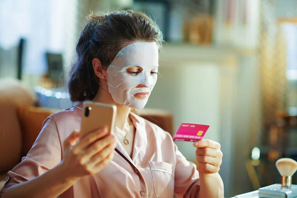 顔に白いシートの顔のマスクを持つパジャマの現代女性は スマートフォンやクレジットカードを保持しています美容化粧品オンラインEコマースサイトで晴れた冬の日に現代の家 — ストック写真