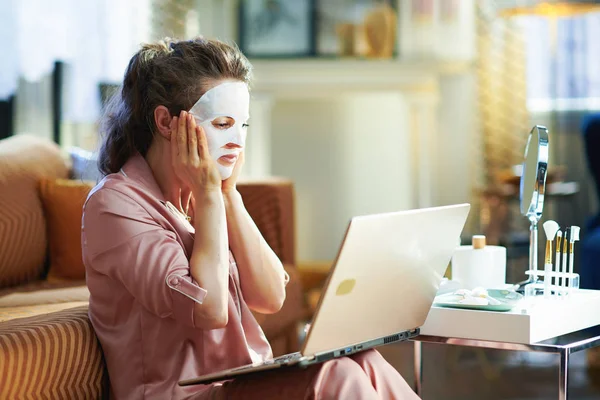 驚くべきエレガントな女性ですパジャマと白いシートの顔のマスクテーブルの近くにバスアメニティとミラーを使用してウェブサイト晴れた冬の日に現代の家のラップトップ上で — ストック写真