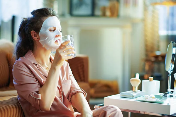 エレガントな女性ですパジャマで顔のマスク顔の上に水を飲みます現代の家で晴れた冬の日 — ストック写真