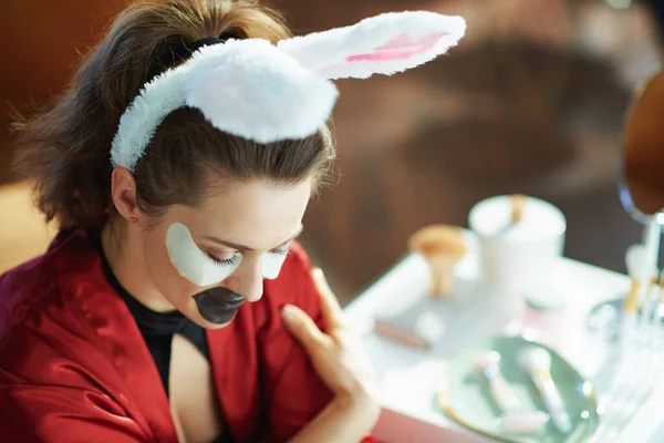 relaxed stylish woman in black body lingerie and red bathrobe with easter bunny ears making cosmetic lip mask and eye mask in the modern house in sunny day.