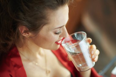 Gündüz vakti modern evde siyah iç çamaşırlı ve kırmızı bornozlu 40 yaşında gülümseyen bir kadın. Bir bardak su içiyor..