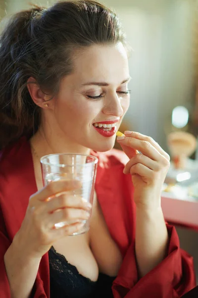 在阳光明媚的日子里 在现代客厅里 身穿黑色内衣 身穿红色浴衣 头戴一杯水的优雅而快乐的女人在吃药 — 图库照片