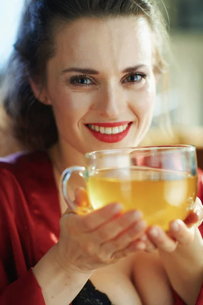 黒体のランジェリーと赤いバスローブで笑顔のトレンディーな女性の肖像画で緑のお茶のカップと現代のリビングルームで晴れた日 — ストック写真