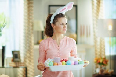 Pembe bluzlu ve paskalya tavşanı kulaklı genç bir kadın güneşli bahar günlerinde modern evdeki mesafeye bakıyor..