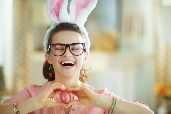40歳の現代主婦の笑顔ピンクのブラウスとイースターのウサギの耳眼鏡で赤いイースターの卵を示す現代のリビングルームで日当たりの良い春の日 — ストック写真