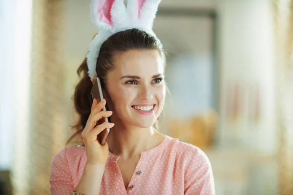ピンクのブラウスを着た若い女性の笑顔と春の晴れた日に現代の家の東のウサギの耳遠くを見てスマートフォンで話す — ストック写真
