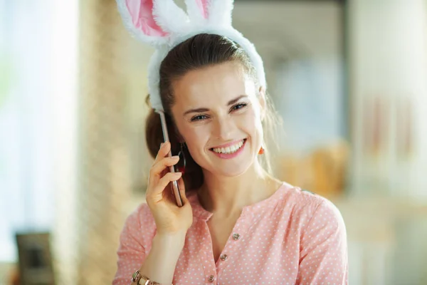 Portrait of smiling modern woman in a pink blouse and easter bunny ears at modern home in sunny spring day using a cell phone.