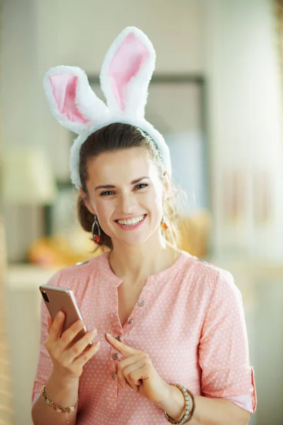 Улыбающаяся Молодая Женщина Розовой Блузке Пасхальных Кроликовых Ушах Смартфоном Использованием — стоковое фото