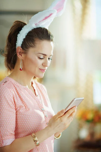 modern female in a pink blouse and easter bunny ears in the modern house in sunny spring day using smartphone for social online connections.