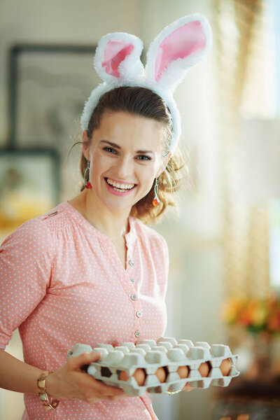 smiling young female in a pink blouse and easter bunny ears in the modern living room in sunny spring day holding big egg carton box.