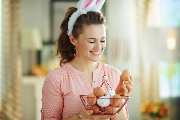 ピンクのブラウスとイースターバニーの耳でエレガントな主婦の笑顔卵とバスケットを保持し 晴れた春の日に現代の家の中で卵を見て — ストック写真