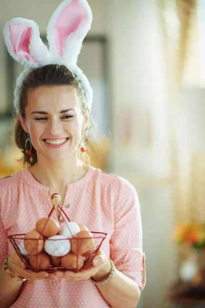 ピンクのブラウスでエレガントな女性を笑顔とイースターバニーの耳を保持バスケットで卵と現代の家で晴れた春の日 — ストック写真