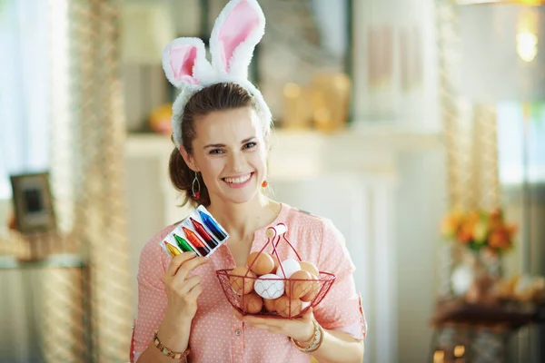 幸せな若い主婦ですピンクのブラウスとイースターバニーの耳ショーバスケットとともに卵と食品着色料で現代のリビングルームで晴れた春の日 — ストック写真