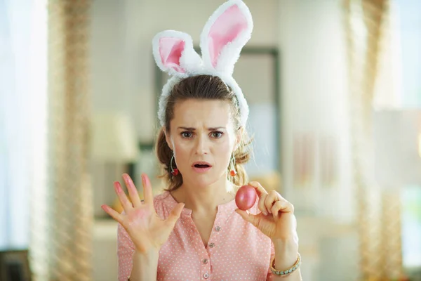 sad modern woman in a pink blouse and easter bunny ears showing hands stained with a red easter egg in the modern living room in sunny spring day.