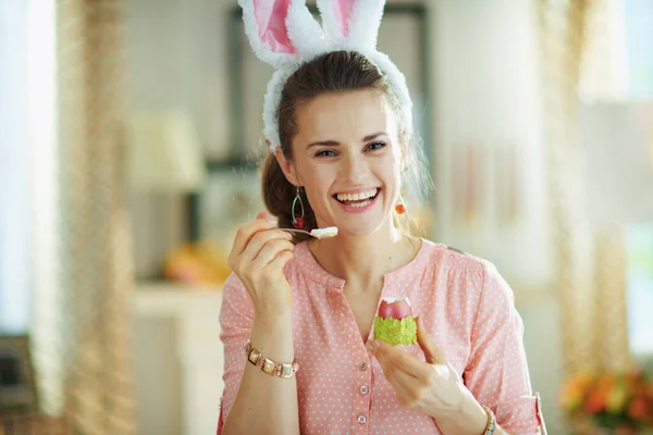 ピンクのブラウスとイースターバニーの耳で幸せなエレガントな女性の肖像画晴れた春の日に現代の家のスプーンで緑の卵カップで赤いイースターの卵を食べる — ストック写真