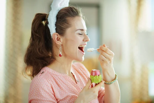 happy young female in a pink blouse and easter bunny ears eating red easter egg in green egg cup with spoon at modern home in sunny spring day.