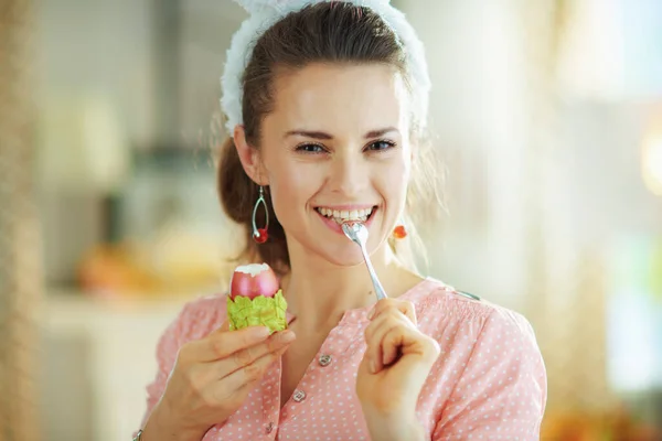 ピンクのブラウスとイースターバニーの耳の若い女性食べる赤いイースターの卵で緑の卵カップとスプーンで現代の家で晴れた春の日 — ストック写真