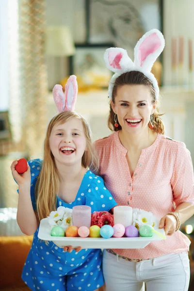 優雅な母親と娘を笑顔でイースターバニー耳ショーイースター装飾プレートでカラフルな卵とろうそくと現代のリビングルームで晴れた春の日 — ストック写真