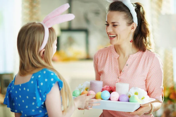 幸せな現代の母親と娘でイースターバニー耳とともにイースター装飾プレートとともにカラフルな卵とキャンドルで現代の家で晴れた春の日 — ストック写真