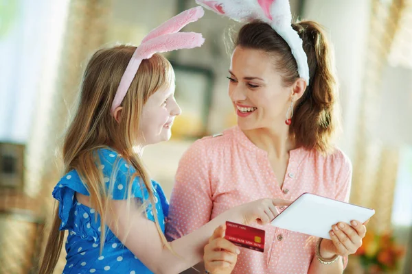 現代の母親と子供を笑顔でイースターバニー耳でクレジットカードオンラインで購入するタブレットPc上で現代の家で晴れた春の日 — ストック写真