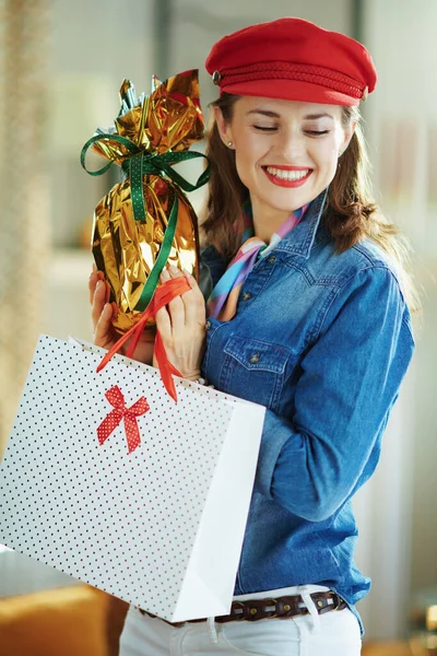 smiling stylish 40 years old woman in a jeans shirt and red hat at modern home in sunny spring day holding wrapped in gold foil big easter egg and white polka dot shopping bag.