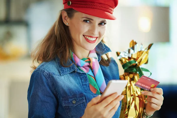 smiling 40 years old woman in a jeans shirt and red hat with credit card and wrapped in gold foil big easter egg buying on internet in the modern living room in sunny spring day.