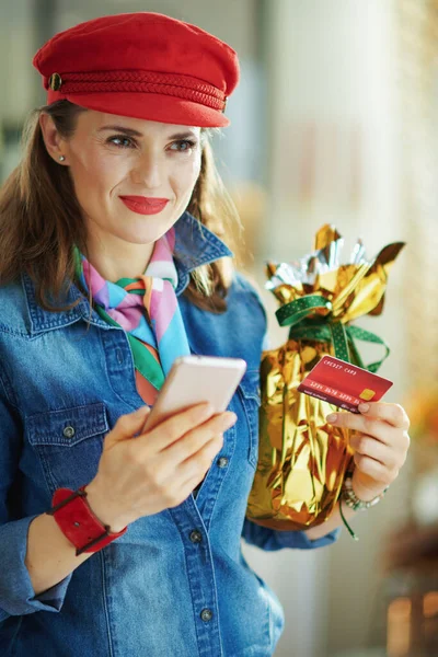 Pensive young woman in a jeans shirt and red hat with credit card and wrapped in gold foil big easter egg making online shopping on e-commerce website in the modern house in sunny spring day.