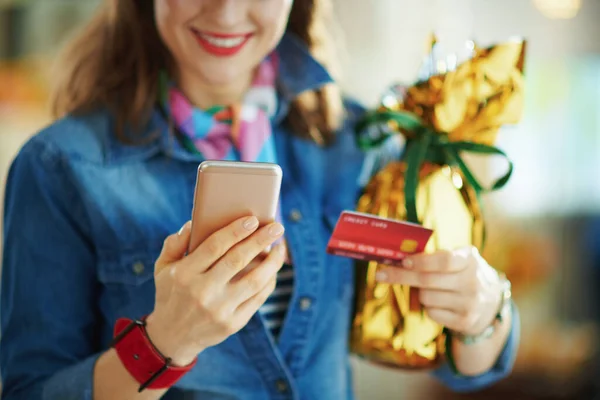 Closeup on happy woman with credit card and wrapped in gold foil big easter egg making online shopping on e-commerce website at home in sunny spring day.