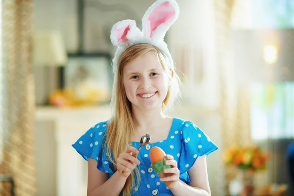 幸せな現代の子供の肖像画でポルカドットブルー全体とイースターバニー耳食べるイースター卵で緑の卵カップでスプーンで現代のリビングルームで晴れた春の日 — ストック写真