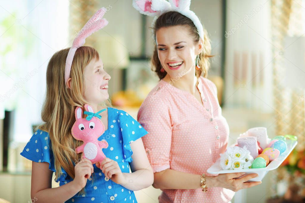 smiling elegant mother and daughter in easter bunny ears with pink rabbit toy and easter decorated plate with colorful eggs and candles in the modern living room in sunny spring day.