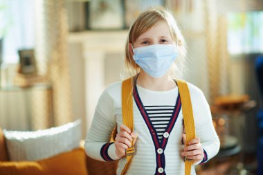 Güneşli bir günde modern bir evde beyaz çizgili tişörtlü, tıbbi maskeli ve sırt çantalı bir çocuk..