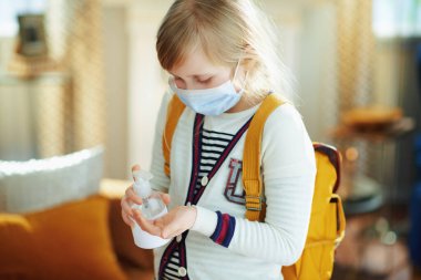 modern child in white striped t-shirt with medical mask and backpack sanitizes hands with a disinfectant at modern home in sunny day. clipart