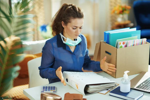 stressed modern woman in blue blouse with medical mask working in temporary home office during the coronavirus epidemic in the modern living room in sunny day.