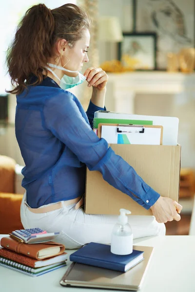 Seen from behind modern woman in blue blouse with medical mask transporting work stuff to a temporary home office during the coronavirus epidemic in the modern living room in sunny day.