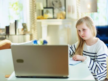 Gündüz modern oturma odasında çizgili süveter giyen genç bir çocuk masada otururken dizüstü bilgisayarla web sitesini kontrol ediyor..