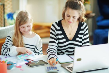 Anne, güneşli bir günde modern oturma odasındaki geçici ev ofisinde çocuk maskesini takarken çalışıyor..