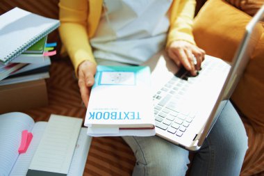 Güneşli bir günde evde dizüstü bilgisayarı ve ders kitabı olan kot pantolonlu ve sarı ceketli bir kadına yaklaş..