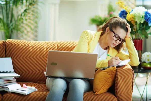 ストレスを感じるエレガントな学習者の女性のジーンズと黄色のジャケット現代のリビングルームで晴れた日の研究オンラインノートパソコン上で — ストック写真