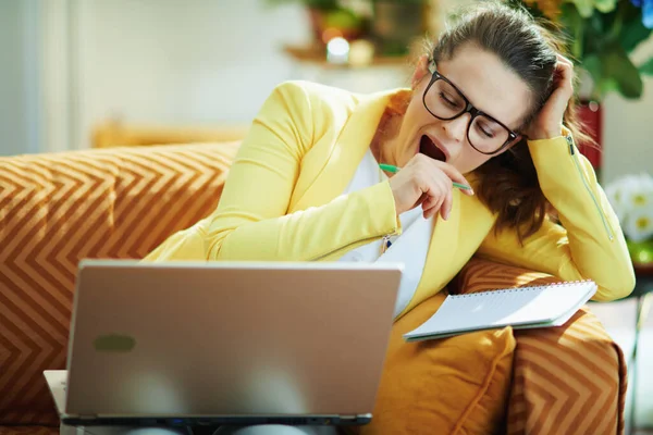 疲れてエレガントな学生の女性でジーンズと黄色のジャケットで現代の家で晴れた日の研究オンライン上のラップトップ — ストック写真