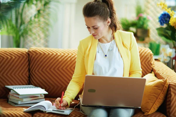 在阳光明媚的日子里 穿着牛仔裤和黄色夹克的中年妇女在现代客厅里使用笔记本电脑上的视频学习网站 — 图库照片