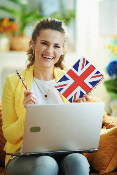 smiling 40 years old woman in jeans and yellow jacket with laptop showing UK flag notebook in the modern living room in sunny day.