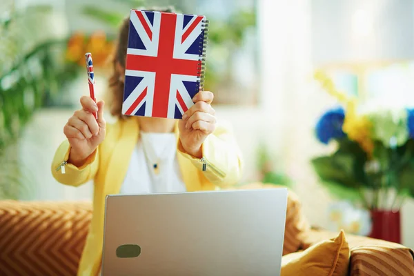 ジーンズの主婦の閉鎖と黄色のジャケットとノートパソコンを持っています晴れた日に自宅で頭の前に英国のフラグノートブック — ストック写真
