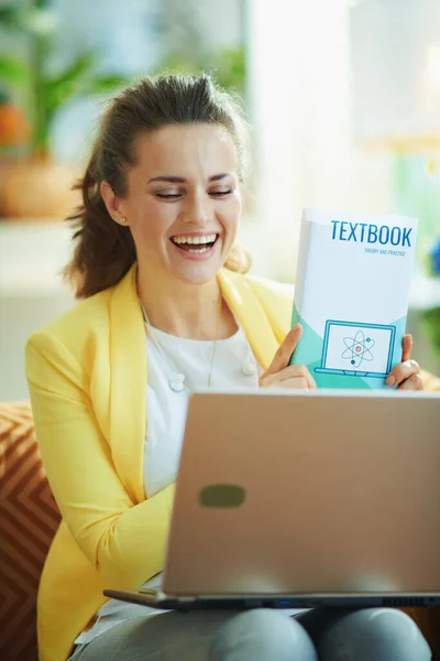 在一个阳光灿烂的日子里 穿着牛仔裤和黄色夹克的快乐的女学生在现代客厅里与一位坐在笔记本电脑上的老师举行的视频会议上展示了课本 — 图库照片