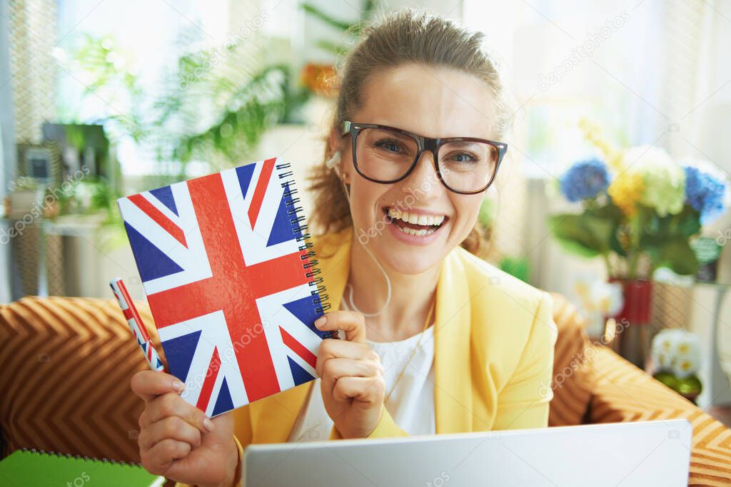 smiling stylish middle age woman in jeans and yellow jacket with laptop showing UK flag notebook at modern home in sunny day.