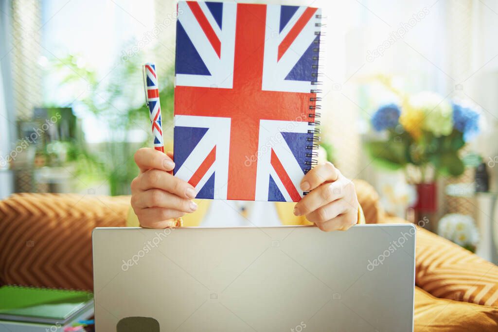 Closeup on learner woman with laptop holding UK flag notebook in the front of head in the house in sunny day.