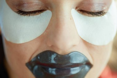 Güneşli bir günde oturma odasında hidrojel dudak maskesi ve göz bandı kullanan bir kadına yakın çekim..