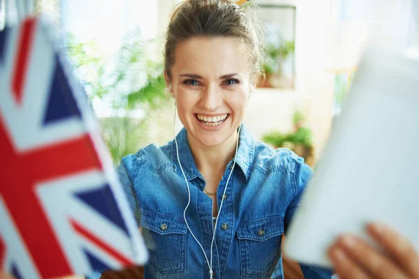 happy modern woman in jeans shirt in the modern house in sunny day showing tablet PC and notebook with English flag