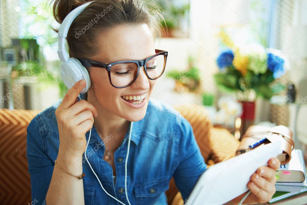 happy middle age housewife in jeans shirt with white headphones and tablet PC study online in the modern house in sunny day.