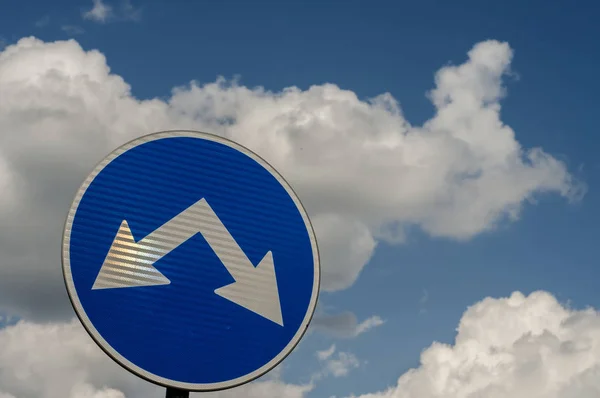 Blue traffic road sign with two arrows for two directions closeup on white clouds blue sky background