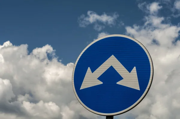 Blue traffic road sign with two arrows for two directions closeup on white clouds blue sky background